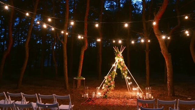 Festive string lights illumination on boho tipi arch decor on outdoor wedding ceremony venue in pine forest at night. Vintage string lights bulb garlands shining above chairs at summer rural wedding.