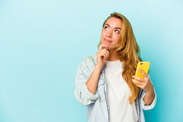 Caucasian woman holding mobile phone isolated on blue background looking sideways with doubtful and skeptical expression.