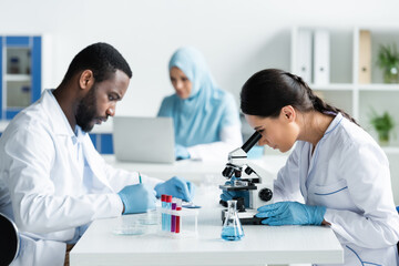 Multiethnic scientists writing on clipboard and working with microscope in lab