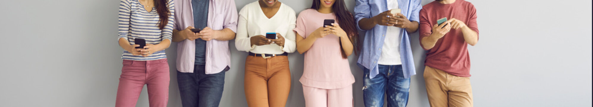 Happy Young People Using Mobile Phones. Diverse Group Of Smiling Zoomers Or Teens Standing In Line Leaning On Wall, Waiting In Queue, Enjoying Online Communication, Connecting To Free Wireless Network