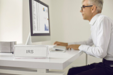 White sign with the inscription IRS on the table on the background of a serious busy senior male tax auditor. Man analyzes the financial performance of the Internal Revenue Service. Audit concept. - obrazy, fototapety, plakaty