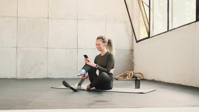 A happy pleased woman with leg prosthesis is taking selfie-photo sitting on the yoga mat in a studio