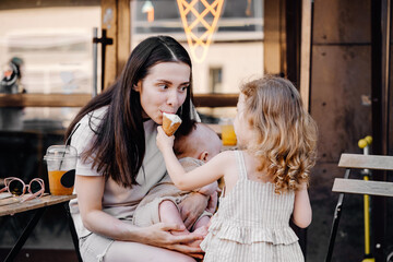 Caucasian toddler girl feeding baby brother boy with ice cream cone. Mother is laughing. Summer family fun outdoors. Sister teasing child with forbidden food