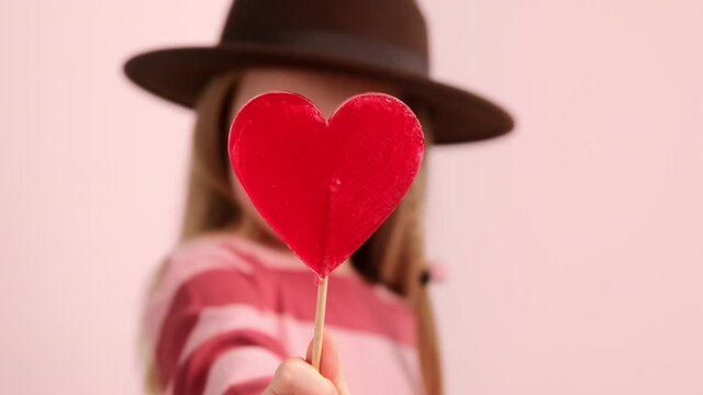 Little girl child in hat holding red heart shape candy, share sends an air kiss. Valentine's day and love sharing concept. Selective blur focus.