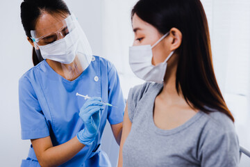 Young Asian woman sitting at doctor's office and getting a flu shot in her arm. Nurse in medical face mask holding syringe and giving female patient modern Covid 19 vaccine injection