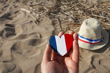 wooden heart in female hands with colors of flag of France blue, white and red on background of golden sand beach with straw hat with ribbon