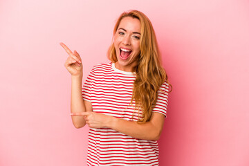 Caucasian blonde woman isolated on pink background pointing with forefingers to a copy space, expressing excitement and desire.