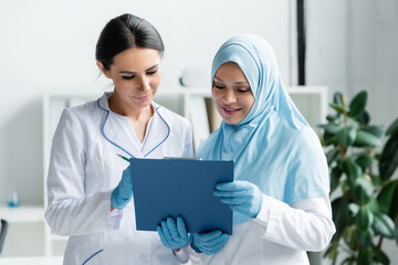 Smiling arabian doctor holding clipboard near colleague