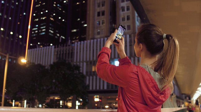Young woman taking photos of buildings on her phone at night, Hong Kong.