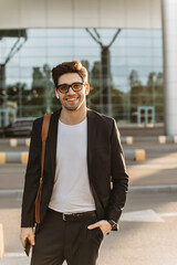 Charming man in black suit, white t-shirt and eyeglasses smiles widely and looks into camera. Brunette guy holds phone and listens to music in headphones outside.