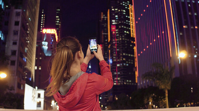 Young woman taking photos of buildings on her phone at night, Hong Kong.