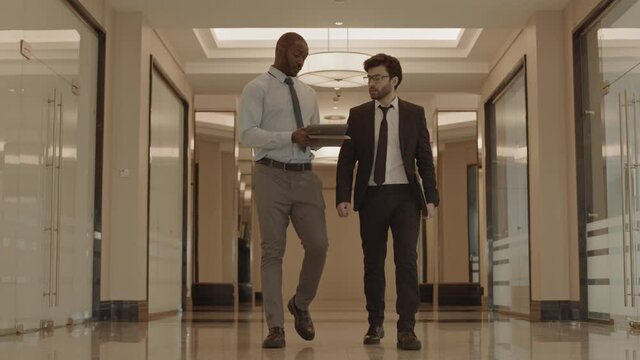 Full tracking of two male coworkers wearing formal clothing, having conversation over tablet computer, walking in hallway of business center