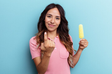 Young latin woman holding ice cream isolated on blue background pointing with finger at you as if inviting come closer.