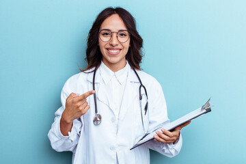 Young doctor mexican woman isolated on blue background smiling and pointing aside, showing something at blank space.