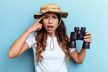 Young mexican woman holding binoculars isolated on blue background showing a disappointment gesture with forefinger.