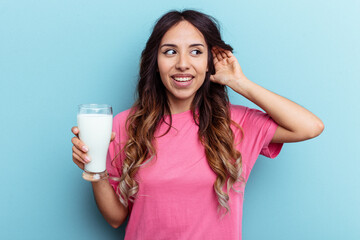 Young mixed race woman holding a glass of milk isolated on blue background trying to listening a gossip.