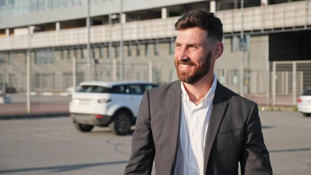 Smiling bearded businessman dressed in classic suit is walking through the parking. Young millennial man waving to friend near white car on the background. Business. Successful people concept.