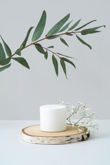 White jar on wooden podium, gypsophila flowers and eucalyptus branch on grey background. Plastic container for advertising cosmetic product on birch saw cut and green leaves. Beauty and spa mockup.
