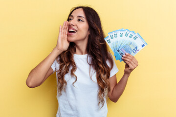 Young mixed race woman holding bills isolated on yellow background shouting and holding palm near...