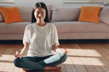 Fotobehang Asian woman doing yoga and zen like meditation lotus pose in casual wear at indoor living room apartment with natural sun light illuminated © primipil