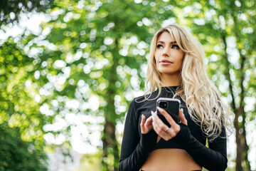 Woman standing hold smart phone typing text message after training outside green park morning. Athletic lady dressed fitness sportwear. Healthy lifestyle, sport recreation. Copy space
