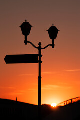 Vertical photography with a silhouette of a vintage street lamps and way sign on the pole, against orange sky at sunset