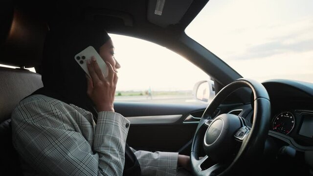 A side view of a happy Arabian businesswoman wearing a national hijab is talking on her smartphone while sitting on the driver's seat