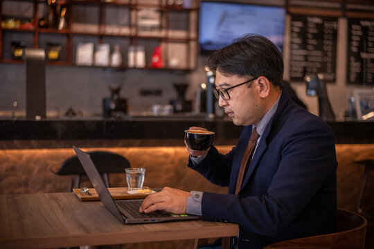Asian businessman wearing suite and eyeglasses drink coffee , typing notebook for work at cafe restaurant. - Powered by Adobe