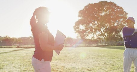 Diverse group of female baseball players and coach on sunny pitch, warming up, stretching arms - Powered by Adobe