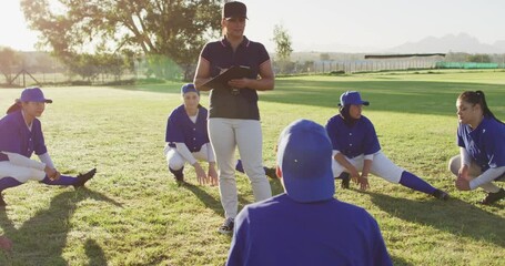 Diverse group of female baseball players with coach, warming up on pitch, squatting, stretching legs - Powered by Adobe