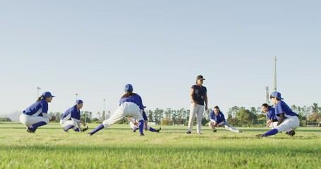 Diverse group of female baseball players and coach warming up on pitch, squatting, stretching legs - Powered by Adobe