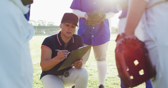 Caucasian female baseball coach squatting writing on clipboard and talking to team on pitch