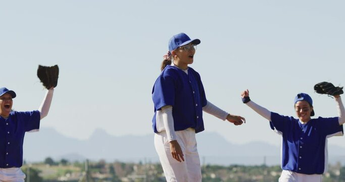 Happy diverse team of female baseball players celebrating after game, embracing and throwing gloves