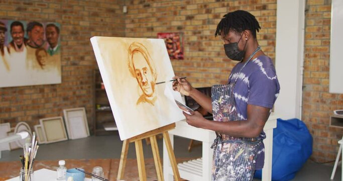African american male painter in face mask painting from photograph on canvas in artist studio