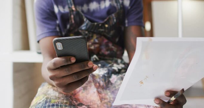 Midsection of african american male painter holding smartphone and photograph in artist studio