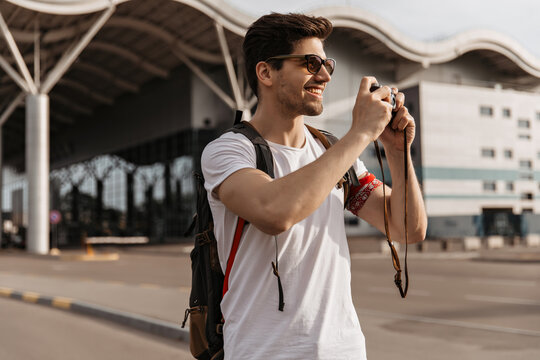 Cheerful guy in sunglasses holds backpack and retro camera. Handsome brunette man in white tee-shirt smiles and takes photos outside.