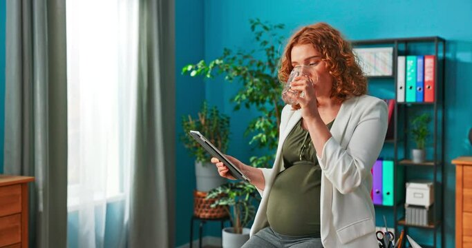 Women With Pregnant Belly Sits At Desk With Tablet In Hand, Drinks Water From Glass, Takes Care Of Hydration, Redhead Can't Wait For Baby To Come Into World, Browses Offers Of Strollers, Clothes