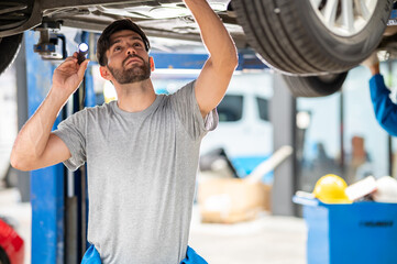 Portrait photo of professional look Caucasian vehicle service technician standing under checking vehicle under body and holding lighting torch in car repair shop for inspect vehicle.	