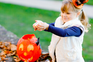Little toddler girl dressed as a witch trick or treating on Halloween. Happy child outdoors, with orange funny hat and pumpkin bag for sweet haunt. Family festival season in october. Outdoor activity