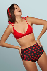 Slim lady is wearing two-piece swimsuit composed of red bra with shoulder straps and black high-waisted panties with floral print. Smiling girl with red headband is posing on the blue background.