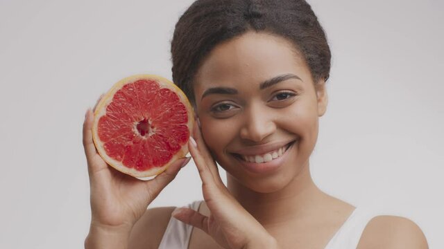 Vitamins for skincare and beauty. Pretty cheerful african american woman holding grapefruit half near her face