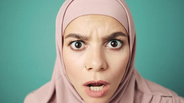 A shocked arabian woman wearing a pink national hijab is looking to the camera standing in the studio isolated over blue background