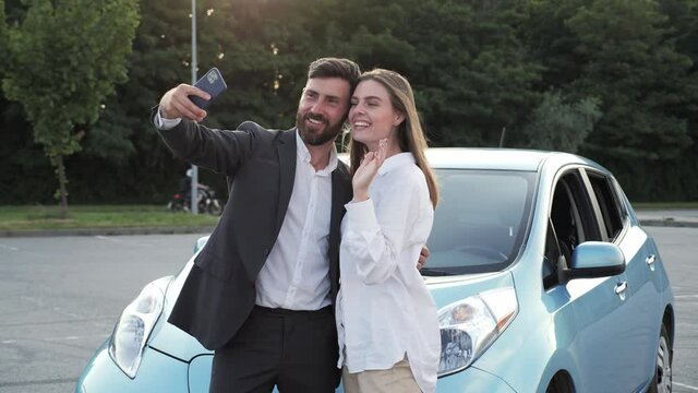 Happy excited loving couple making selfie on the smartphone after purchasing standing near their new electric car feeling expressing gladness. Happy customers dreams come true lifestyle concept.