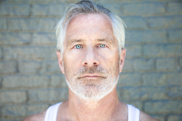 Fit Man with grey hair and beard blue eyes wearing white tank top facing camera with grey brick backdrop