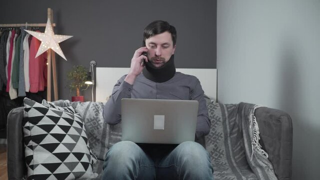 Man in neck brace cervical collar working from home teleworking. Injured male in foam cervical collar working at home talking on phone with laptop sitting on the couch and getting sick from neck pain.