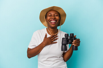 Young african american woman holding binoculars isolated on blue background laughs out loudly keeping hand on chest.