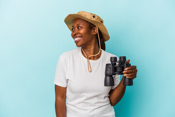 Young african american woman holding binoculars isolated on blue background looks aside smiling,...