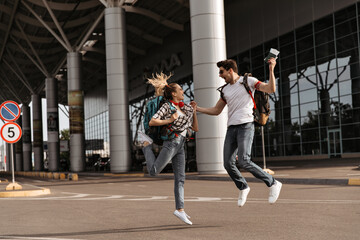 Happy blonde woman and brunette man jumps with backpacks near airport. Joyful travelers in sunglasses poses in good mood and moves.