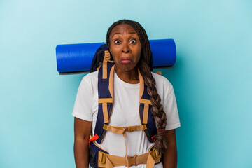 Young african american hiker woman isolated on blue background shrugs shoulders and open eyes...