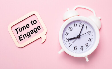 On a delicate pink background, a white alarm clock and a wooden frame with the text TIME TO ENGAGE